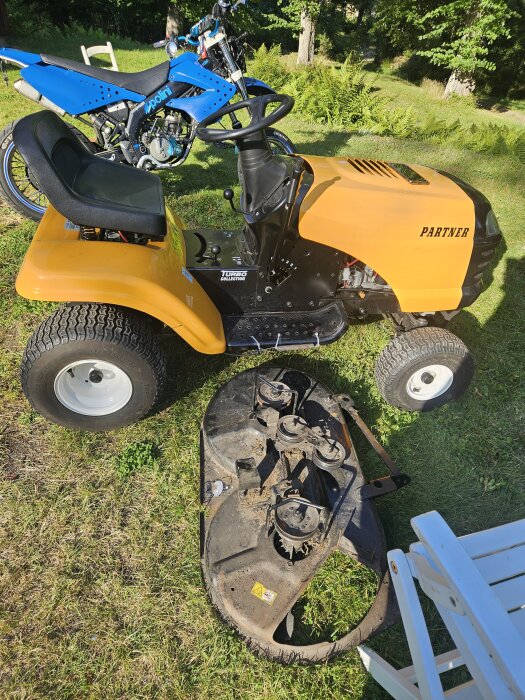Gul åkgräsklippare Partner Electrolux P1192 med ett fristående klippaggregat på marken och en blå motorcykel i bakgrunden.