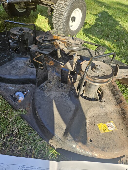 Klippaggregat från en åkgräsklippare ligger på gräsmatta, rostigt och delvis trasigt. Ett papper med instruktioner syns i bildens nederkant.