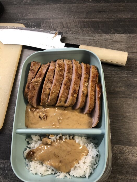 Frukost- och lunchlåda med skivad köttfärslimpa och sås i ena facket, och ris med sås i det andra, på ett köksbord bredvid en kniv.