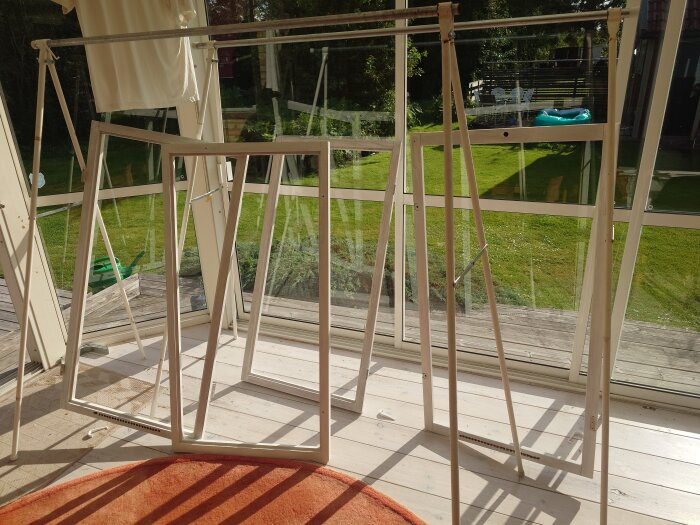 Fönsterkarmar står på tork inomhus i solen, upphängda på en ställning framför stora fönster, med en grön gräsmatta synlig utanför.