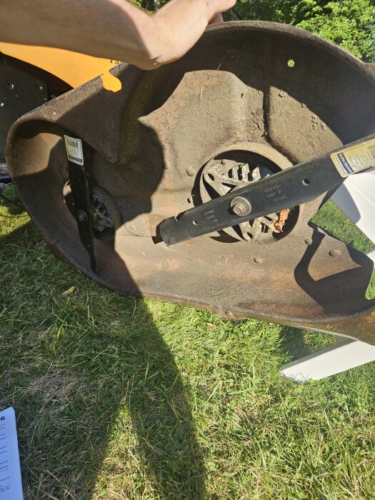 Rostigt klippaggregat på gräsmatta, saknande delar och avbrutet fäste syns; en hand håller i det, manual på marken bredvid.