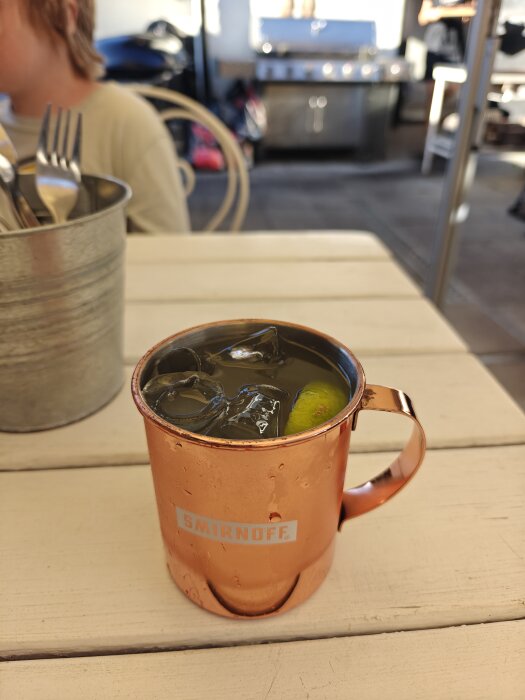 Kopparmugg med Moscow mule på bord, bredvid metallhink med bestick, i en utomhusmiljö med en grill i bakgrunden.