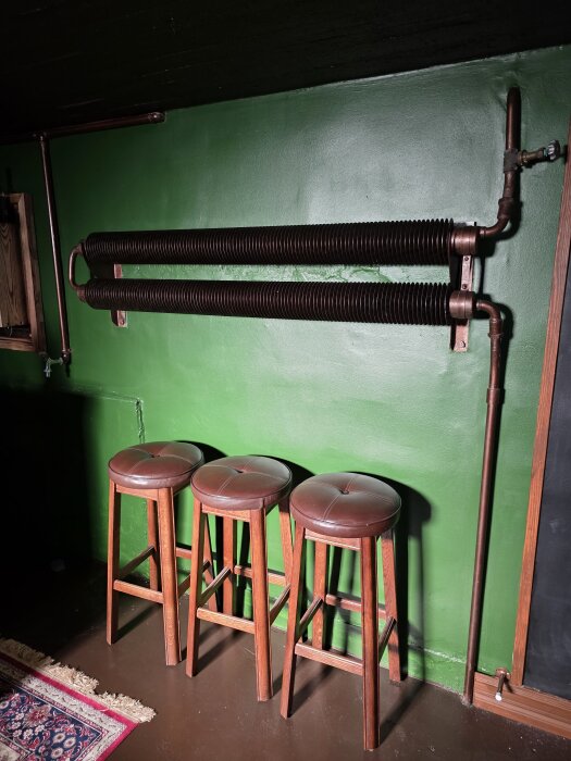 Gamla radiatorer på grön vägg med kopparrör, tre höga pallar framför.