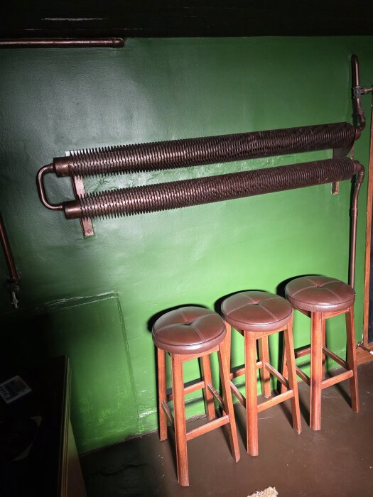 Gamla radiatorrör på grön vägg med tre bruna barstolar framför, i en källare.