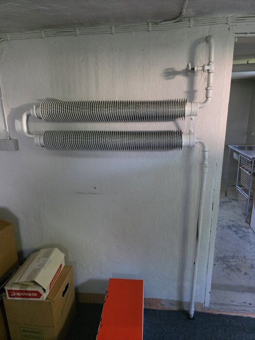 Gamla flänsradiatorer monterade på en vit vägg i en källare, omgivna av rör och några kartonger på golvet.