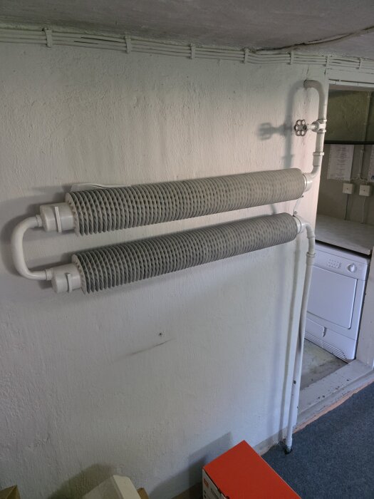 Gamla vita radiatorer med ribbad konstruktion monterade på en vit vägg i en källare, med röranslutningar och en ventil.