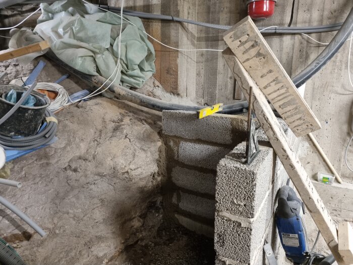 Byggarbetsplats med gjutformar och block, verktyg och kablar på marken, samt en vattenpass placerad på en stapel av betongblock.