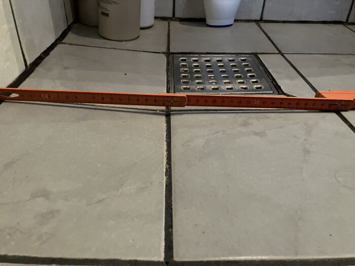 Golvet i ett badrum med en fogmassa nära en brunn och en fällbar mätsticka som ligger på kaklet.