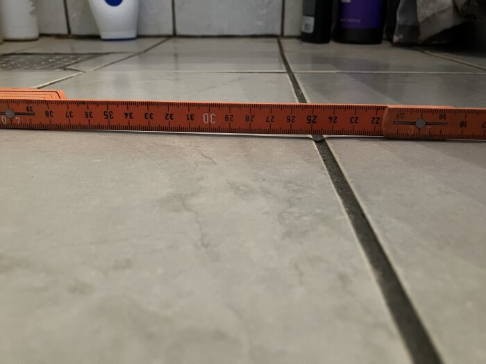 Orange linjal placerad längs ett badrumsgolv för att visa nivåskillnad mellan plattorna. Lindrig nedsjunkning synlig i foglinjen.