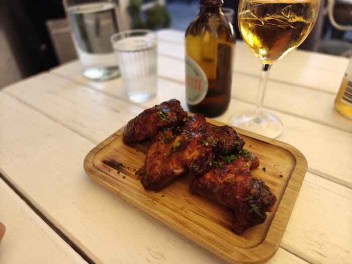 Grillade kycklingvingar på en träbricka, omgiven av ett glas öl och en flaska på ett vitt bord.