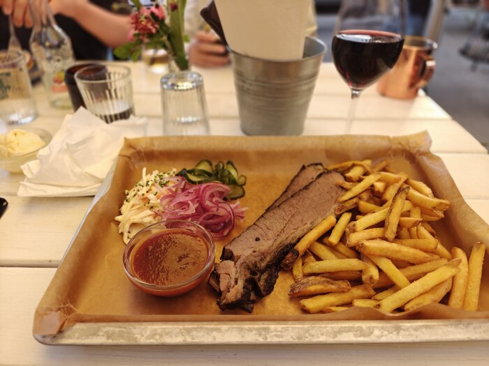Tallrik med bbq-kött, pommes frites, inlagd rödlök, gurka, coleslaw och sås på en vit bordsduk med vinglas och dukar i bakgrunden.