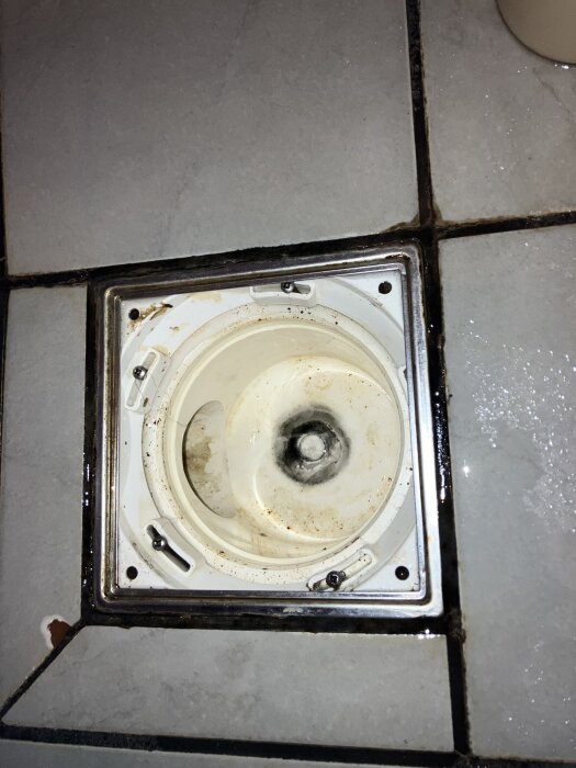 Avlopsbrunn på kaklat golv med smuts och avlagringar, visar bristande regling runt brunnen.