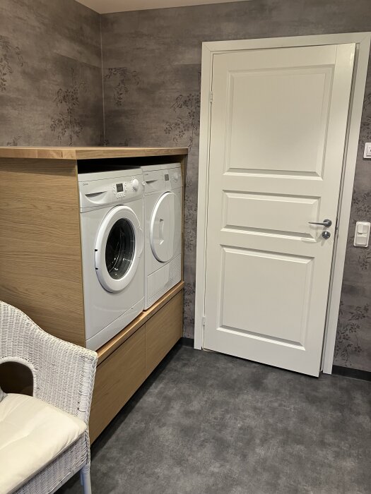 Badrum med tvättmaskin och torktumlare upphöjda för bättre arbetshöjd, placerade bredvid en vit dörr, gråblommigt tapetmönster i bakgrunden.