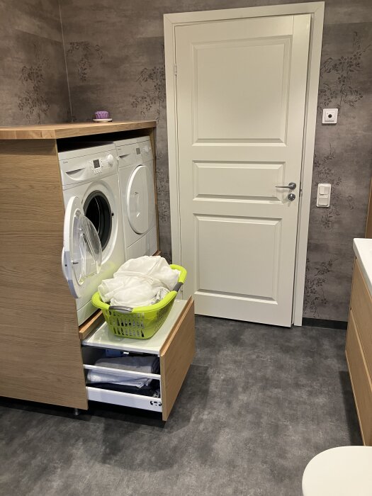 Badrum med tvättmaskin och torktumlare i träskåp, med en tvättkorg på utdragbar hylla, grått golv och blommigt tapetmönster.