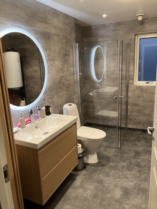 Modernt badrum med spegel med belysning, träkommod, toalett och duschhörna med glasväggar, grå väggar med mönster och grått golv.