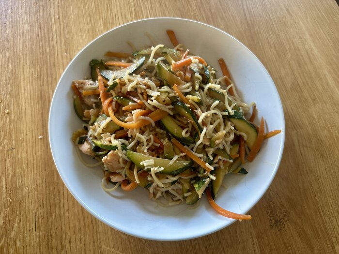 Tallrik med nudlar, grönsaker som morötter och zucchini samt kyckling, på ett träbord.