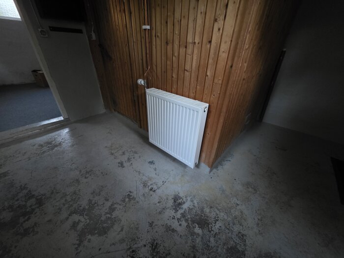 Radiator vid träpanelvägg i källare med ett betonggolv och ett öppet rum i bakgrunden.