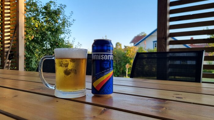 En ölglas fylld med skummande öl och en ölburk står på ett träbord utomhus med en trädgård i bakgrunden.