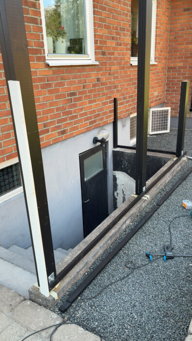 Byggprojekt vid källartrappa med nya stolpskor och reglar monterade på tegelvägg. Verktyg och material syns vid grusgång.