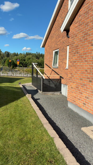 Tegelhus med ny källartrappa i metall med tak av plåt och träreglar, omgiven av grus och gräsmatta under blå himmel.