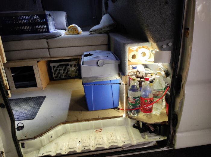 Interiör av en skåpbil med LED-belysning, kylbox, plastflaskor, filtrulle, och vikta filtar på en soffa.