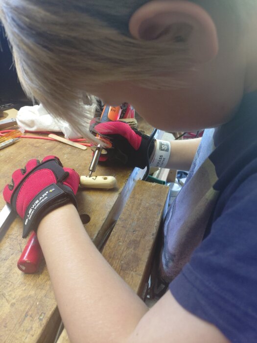 Barn bränner initialer på ett eldstål som present med en brännpenna, iklädd röda handskar vid en arbetsbänk.