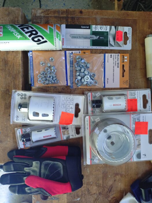 Verktyg och byggtillbehör från Jula, inklusive hålborrset, muttrar, arbetshandskar och energidryck, på ett arbetsbord. Rabattetiketter synliga.