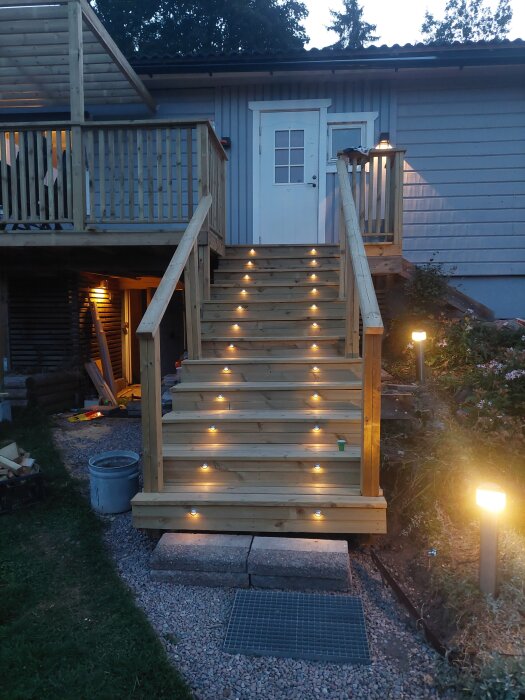 Trätrappa med installerade spottar i varje steg, omgiven av ljus från ytterbelysning och leder upp till en altan med ett vitt dörrparti.