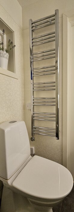 Handdukstork installerad ovanför toalett i ett badrum, med vitmålad vägg och hylla med blomma i bakgrunden.