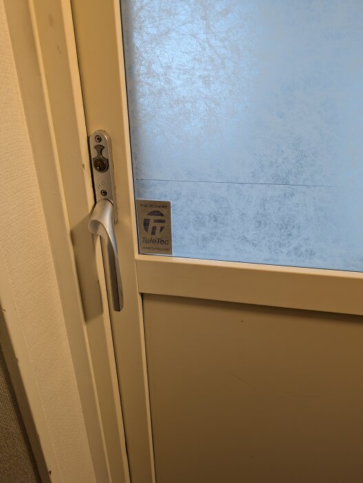 Dörr med ett vitt handtag och låscylinder, delvis frostat glas med "TeleTec" märke. Kan bytas mot elektroniska handtag för låsning från båda sidor.