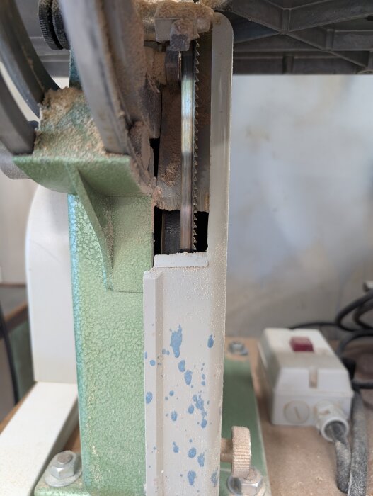 Närbild av begagnad bandsåg med dammsugarmunstycke monterat, täckt av fint damm i verkstadsmiljö.