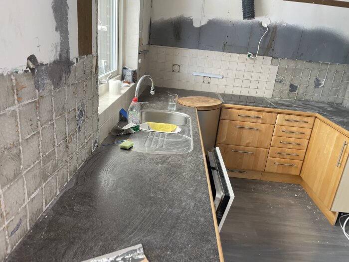 Kök under renovering med delvis borttagen kakel, diskbänk med rengöringsmedel och svampar, och träluckor.