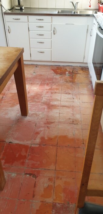 Slitet köksgolv i tegelröd nyans med märken och fläckar, omgiven av vita skåp och en diskbänk.