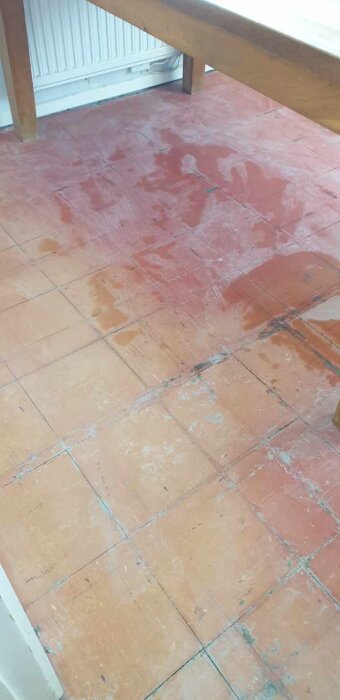 Slitet golv med fyrkantiga plattor i ett rum, synlig radiator och träbord ovanför.