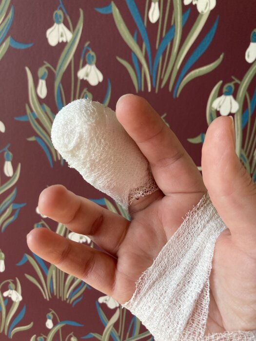 Hand med bandagerat långfinger efter ganglionoperation, mot bakgrund av mönstrad tapet med vita blommor.