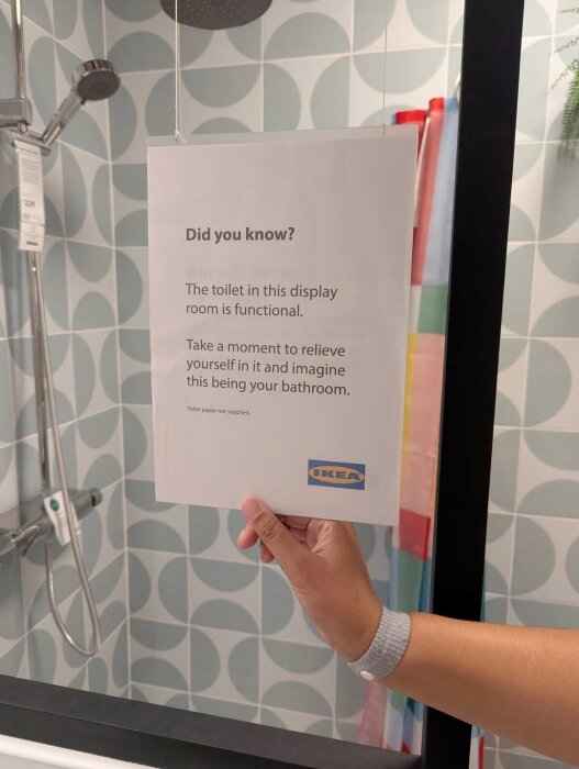 Person håller en skylt på IKEA med text om en fungerande toalett i visningsrummet, omgiven av ett modernt badrumsinredning.