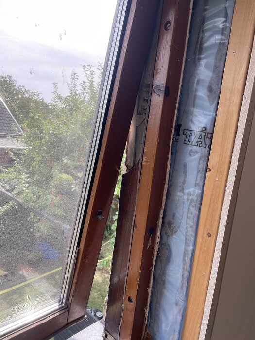 Gammalt träfönster på andra våningen, delvis borttaget inifrån. Fönsterramen och tätningen syns tydligt, med utsikt över grönska utanför.