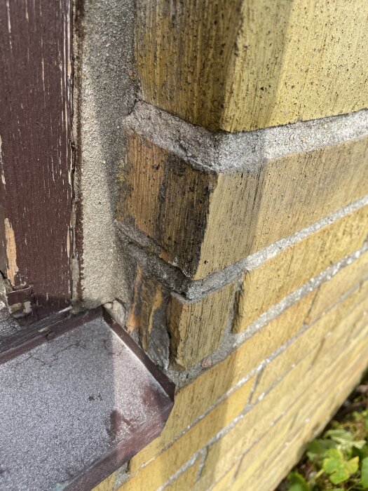 Skadat fönsterparti med synliga tegelväggar och fog, nära en brun fönsterkarm, ovanpå en bottenplåt, som visar väderpåverkan.