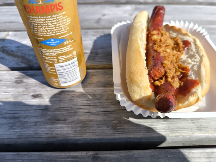 Korv med bröd och rostad lök pÄ en bÀnk utomhus, bredvid en Champis-dryck.