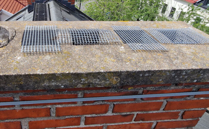 Skorsten med fyra ventilationsgaller, där ett är snett och exponerar rökgången. Platt takomgivning, rött tegel och gröna träd syns.