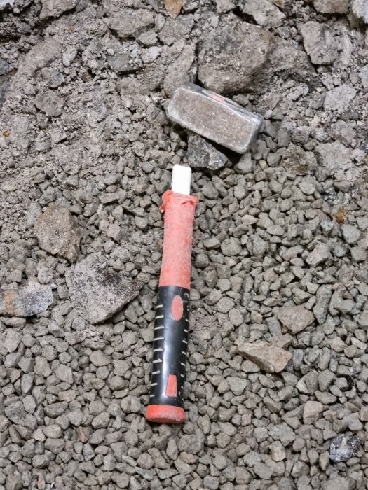 Hammare med rött handtag på grusig mark omgiven av små stenar.
