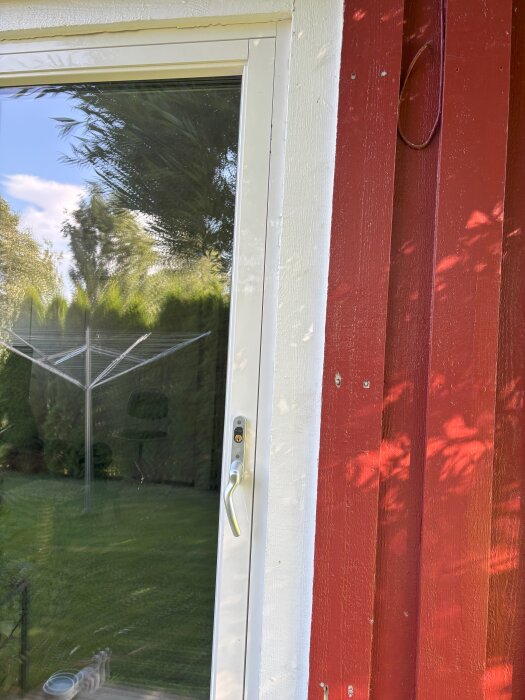 Glasdörr med vitt gångjärn och löst dörrhandtag på en röd trävägg med grön gräsmatta i bakgrunden.