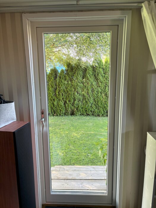 Helglasad utåt öppnande dörr med högt gräs och buskar utanför, dörrhandtaget verkar löst. Dörrbladet hänger något, med öppen glipa längst ner.