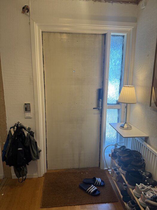Ytterdörr med synliga skruvhål och en skoställning bredvid, i en hall med parkettgolv. Lampan står på en liten hylla ovan radiatorn.