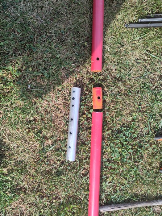 Röda och silverfärgade rör på gräsmattan med borrade hål för låsning, ett del av ett reparationsprojekt för ett trasigt mål.