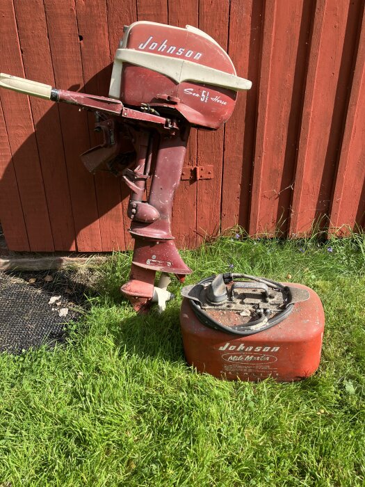 Gammal Johnson Sea Horse utombordsmotor lutad mot en röd vägg, bredvid en bensintank på gräsmattan.