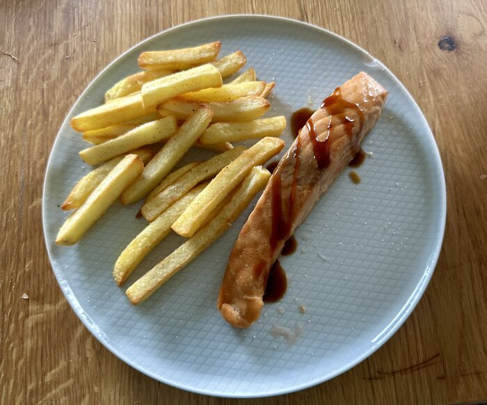Tallrik med pommes frites och lax med sÄs pÄ trÀbord.
