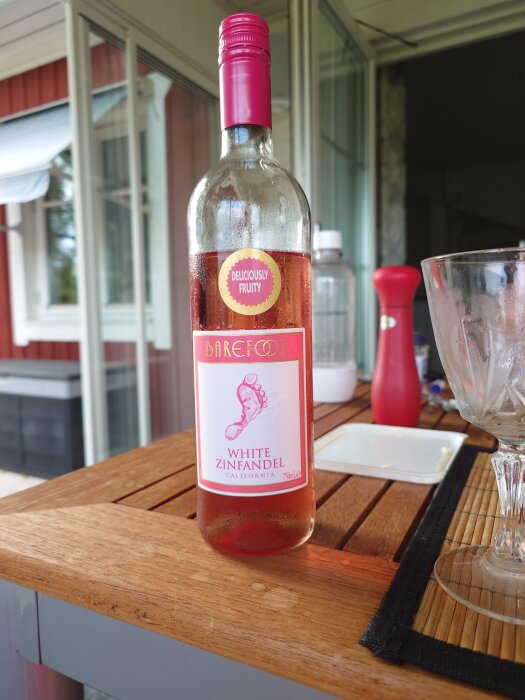 Flaska med rosévin, Barefoot White Zinfandel, står på ett bord utomhus med glas och kryddkvarn i bakgrunden.