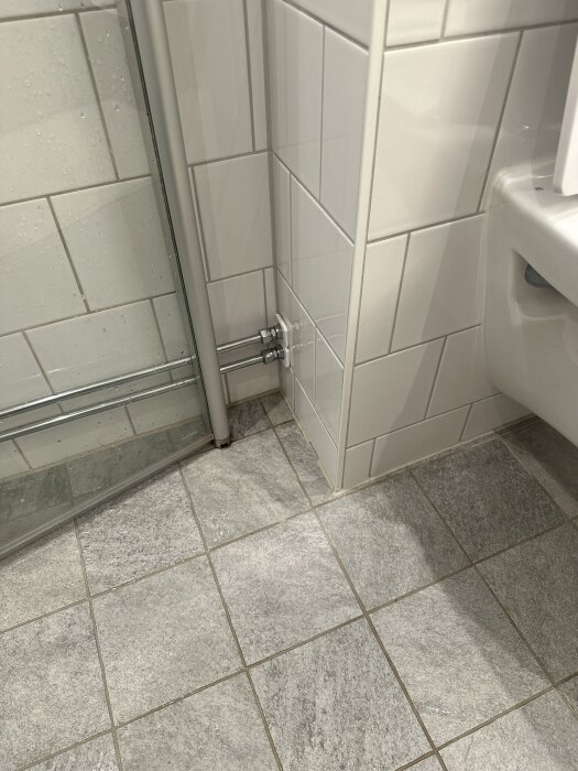 Vitt kaklat badrum med grått klinkergolv, dusch, vägghängd toalett och synligt hål i silikonfogen mellan duschen och toaletten.