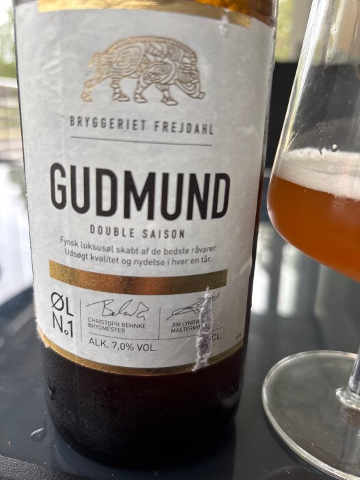 Flaska Gudmund Double Saison från Bryggeriet Frejdahl och ett glas öl på ett bord. Etiketten visar alkoholhalten 7,0%.